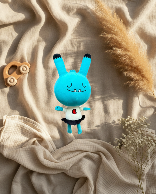 Peluche monstre d’amour turquoise, yeux fermés, cœur rouge sur bavoir blanc, jambes noires.